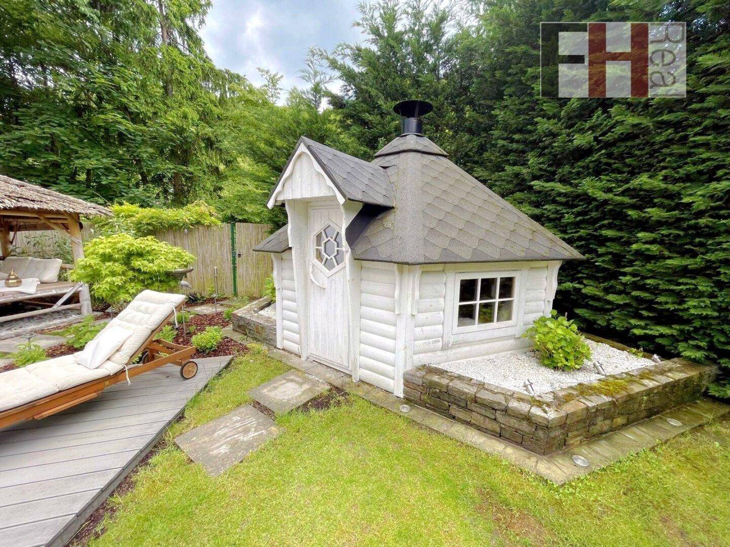 Charmantes weißes Gartenhaus mit dunklem Dach und kleiner Steinumrandung im grünen Garten.