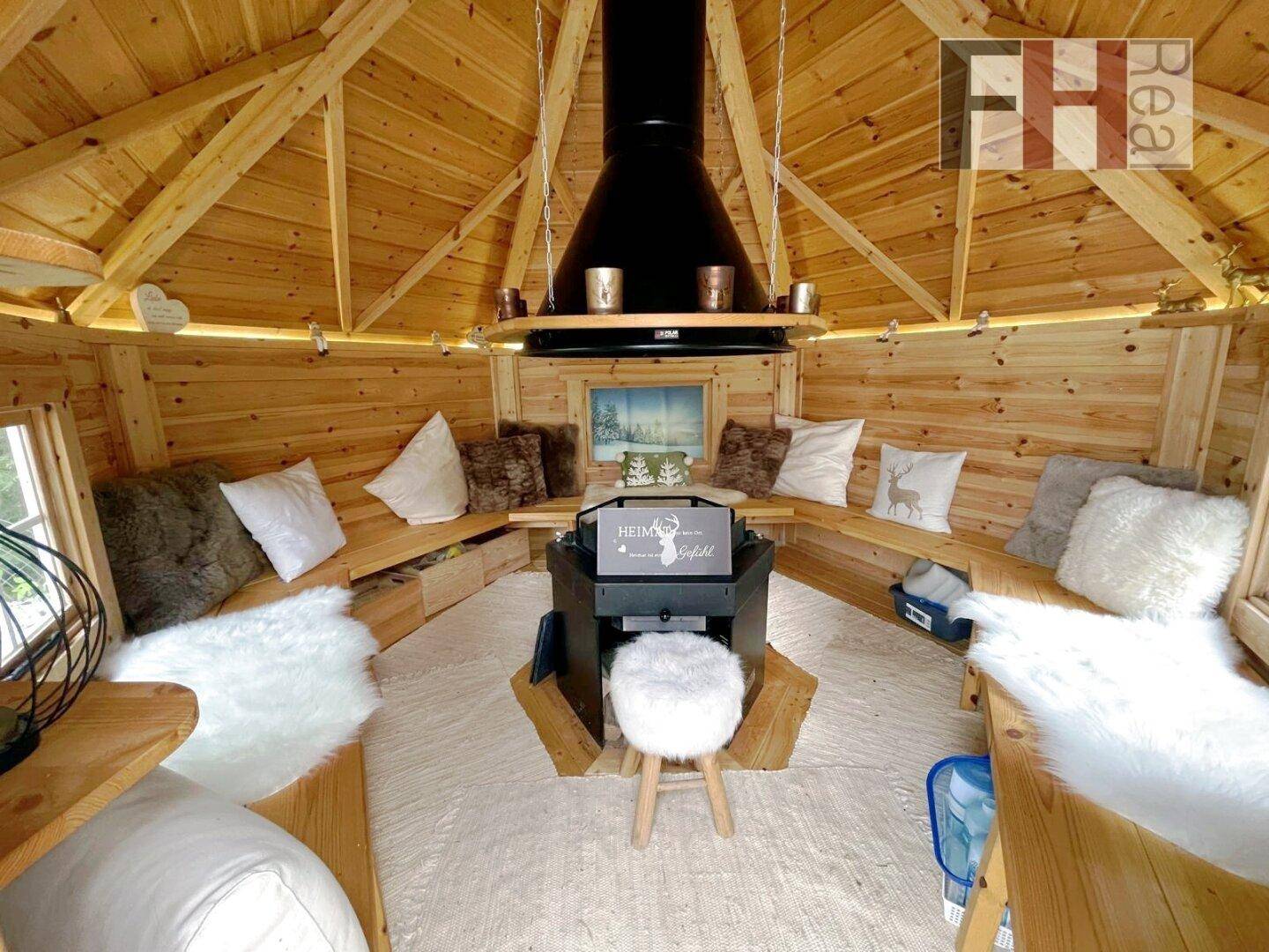 Gemütlicher Innenraum einer Grillhütte mit Holzbänken, Fellen und zentralem Grillbereich.