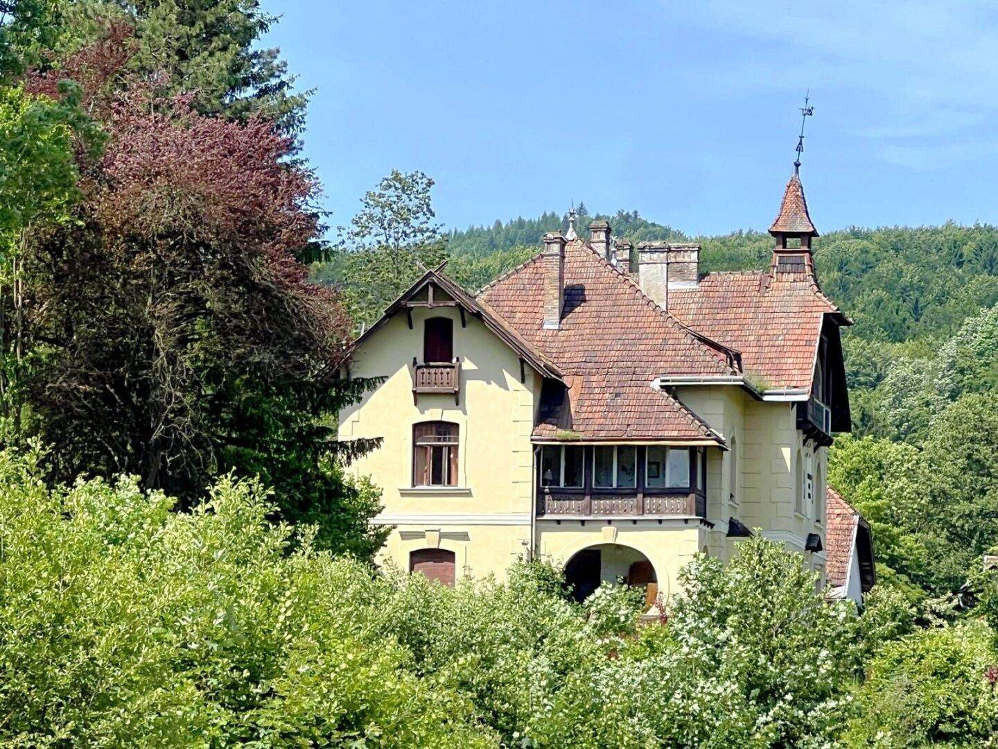 Seitenansicht des historischen Gebäudes mit gelber Fassade und roten Dachziegeln, umgeben von Bäumen.