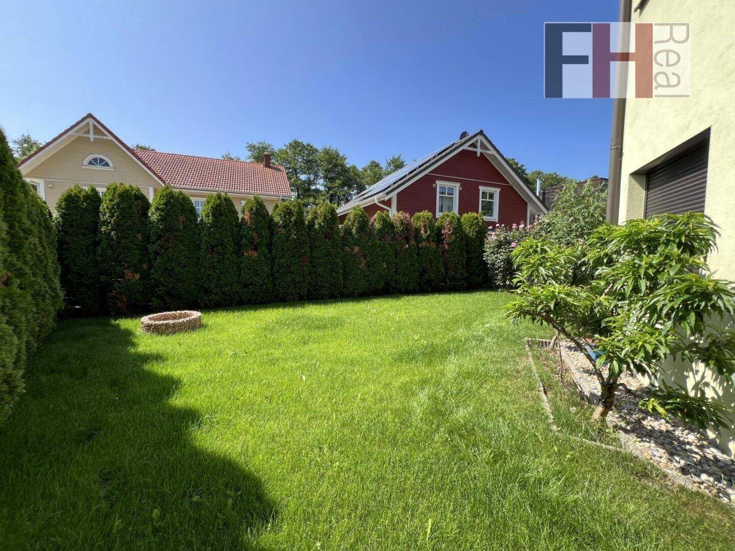 Weitläufiger Rasenbereich mit Feuerstelle, umgeben von einer Hecke und Blick auf benachbarte Häuser.