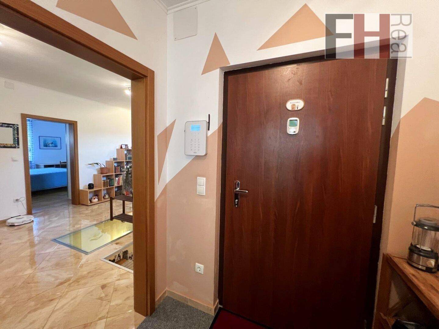 Flur mit hellen Fliesen, Sicherheitssystem an der Wand und Zugang zu weiteren Räumen.