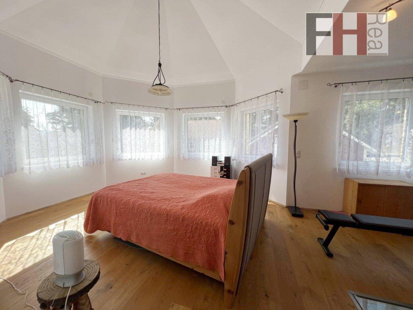 Helles Schlafzimmer mit großem Bett, Holzfußboden und zahlreichen Fenstern, die den Raum erhellen.