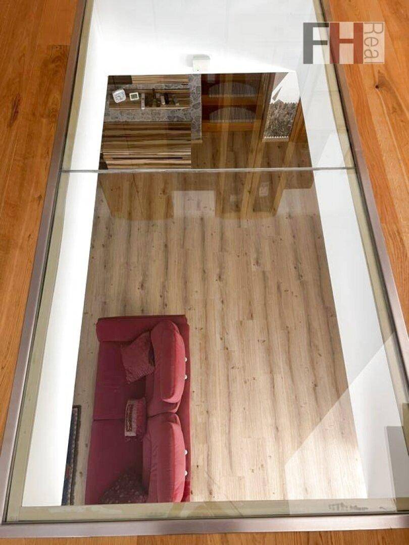 Blick durch ein Glasbodenelement auf das darunterliegende Wohnzimmer mit rotem Sofa.