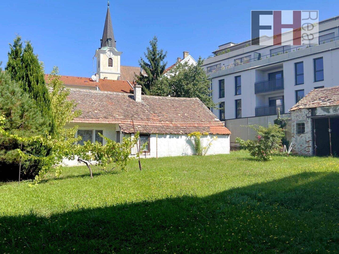 Weitläufiger Garten mit altem Haus, umgeben von Grünflächen und einem Kirchturm im Hintergrund.