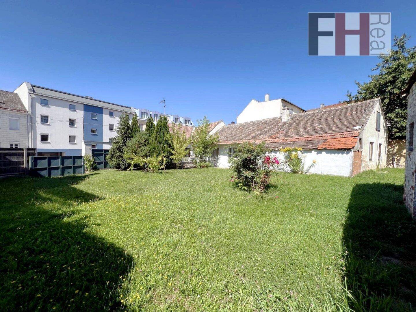 Große Rasenfläche eines Gartens mit einem älteren Haus und angrenzenden Gebäuden unter blauem Himmel.