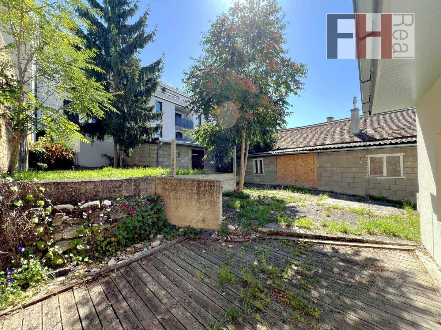 Hölzerne Terrasse mit Pflanzen und Blick auf ein Nebengebäude sowie moderne Architektur im Hintergrund.