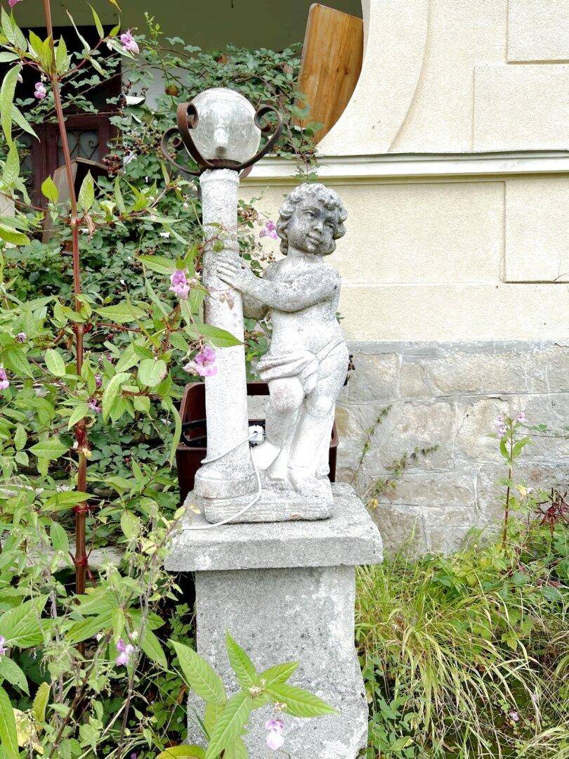Steinstatue eines Putten im Garten, umgeben von grünen Pflanzen und kleinen Blüten.