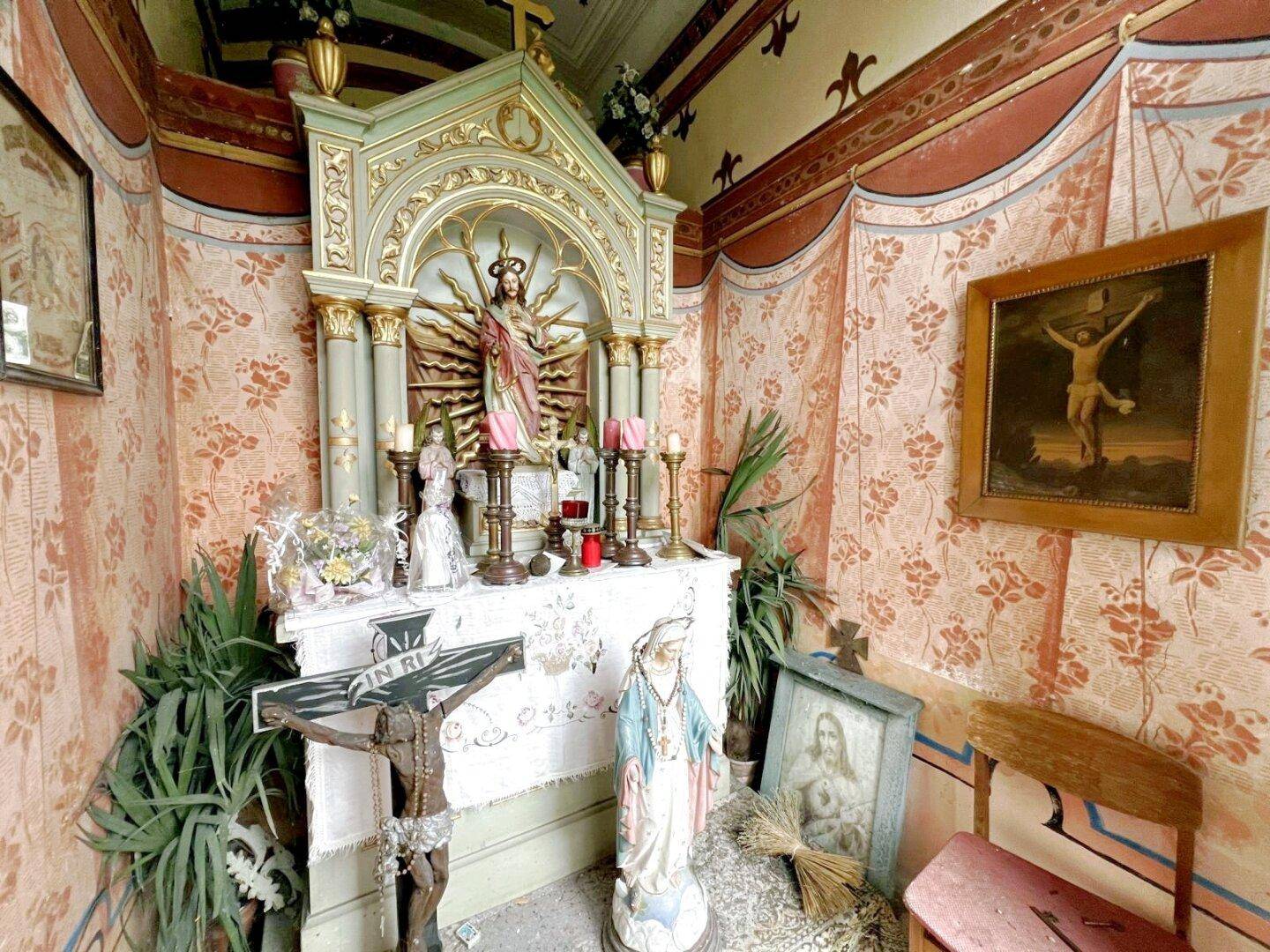 Kleiner Andachtsraum mit religiösen Statuen, Gemälden und floraler Tapete.