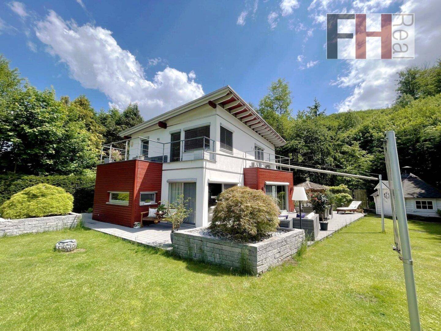 Modernes Einfamilienhaus mit weißer Fassade, rotem Akzent, großer Terrasse und gepflegtem Garten in Hanglage.