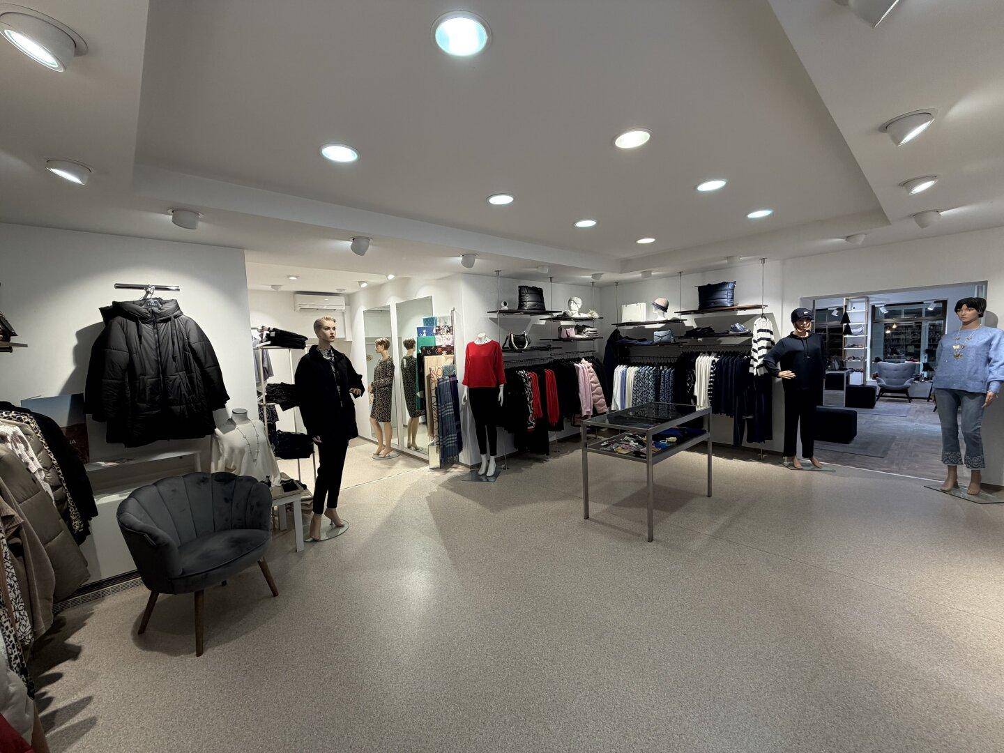 Übersichtlicher Verkaufsraum einer Boutique mit Kleiderständern, einem Sessel und einem Präsentationstisch.