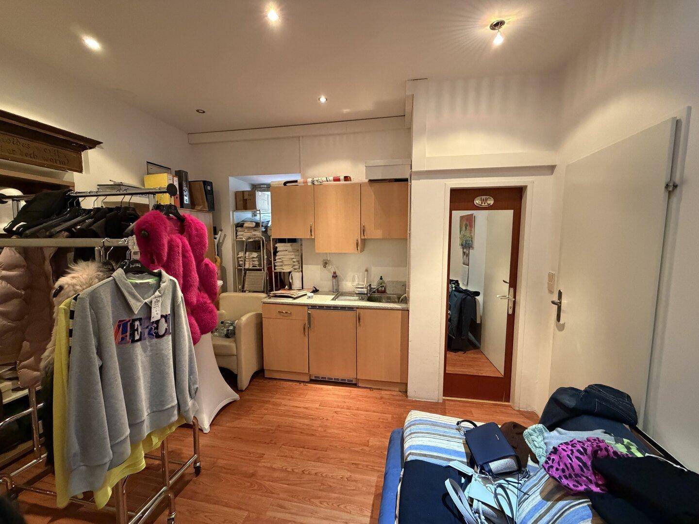 Hinterzimmer mit kleiner Küchenzeile, Holzfußboden und Kleiderständern, dient als Lager- und Pausenbereich.