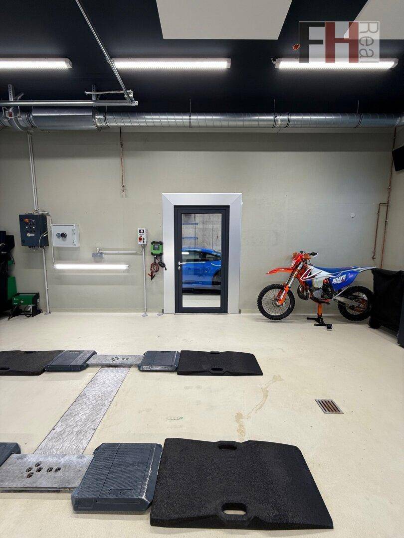 Geräumige Werkstatt mit einem Motorrad, einer Tür, die ein blaues Auto reflektiert, und industriellen Deckeninstallationen.