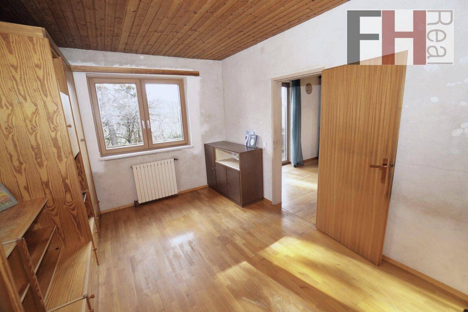 Helles Zimmer mit Holzboden, Holzdecke und Fenster mit Blick auf die Umgebung.