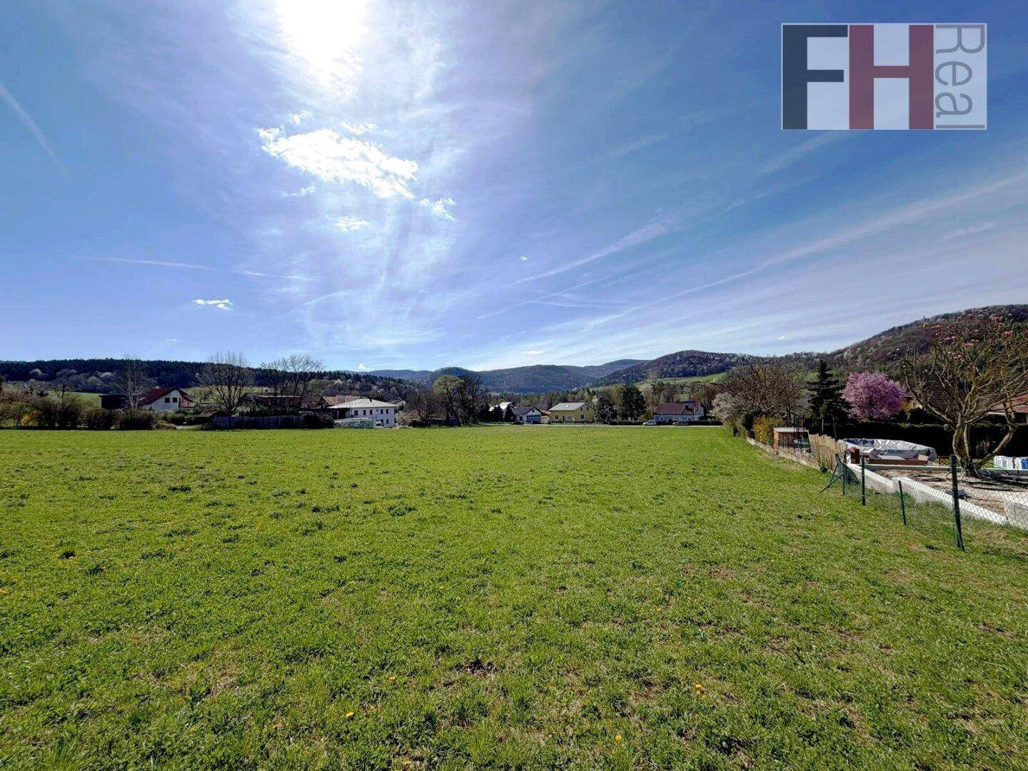 Sonniges grünes Feld mit ländlicher Bebauung und bewaldeten Hügeln am Horizont.