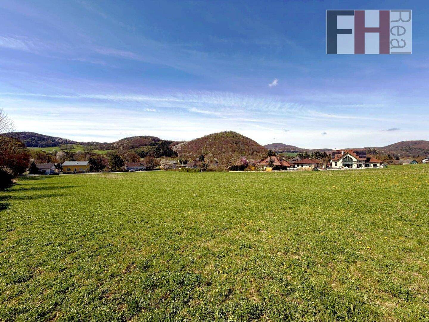 Idyllisches grünes Feld mit ländlicher Bebauung und einer malerischen Hügellandschaft im Hintergrund.