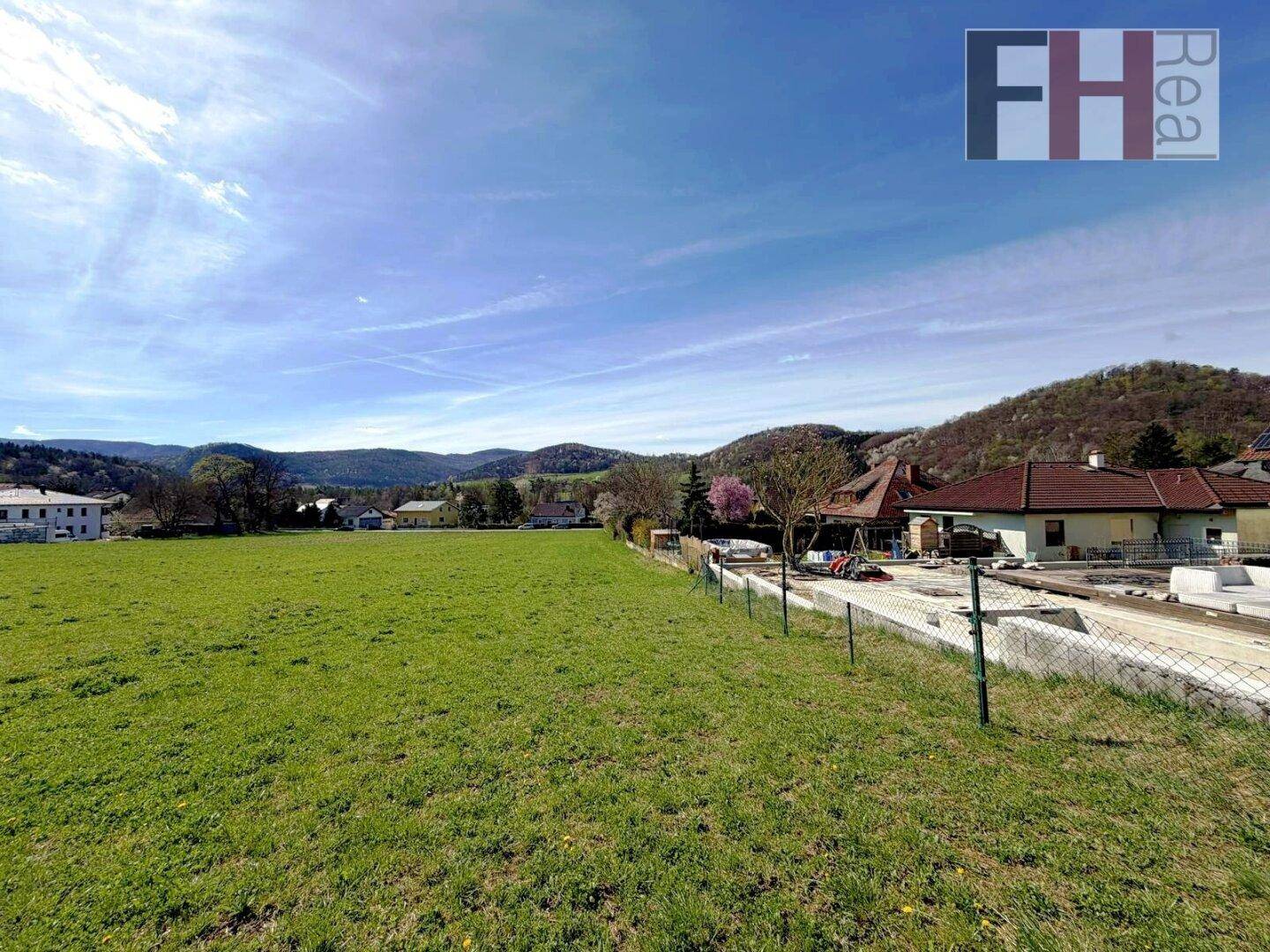 Sonniges grünes Feld mit Blick auf Häuser, Hügel und eine Baustelle am Rande des Grundstücks.