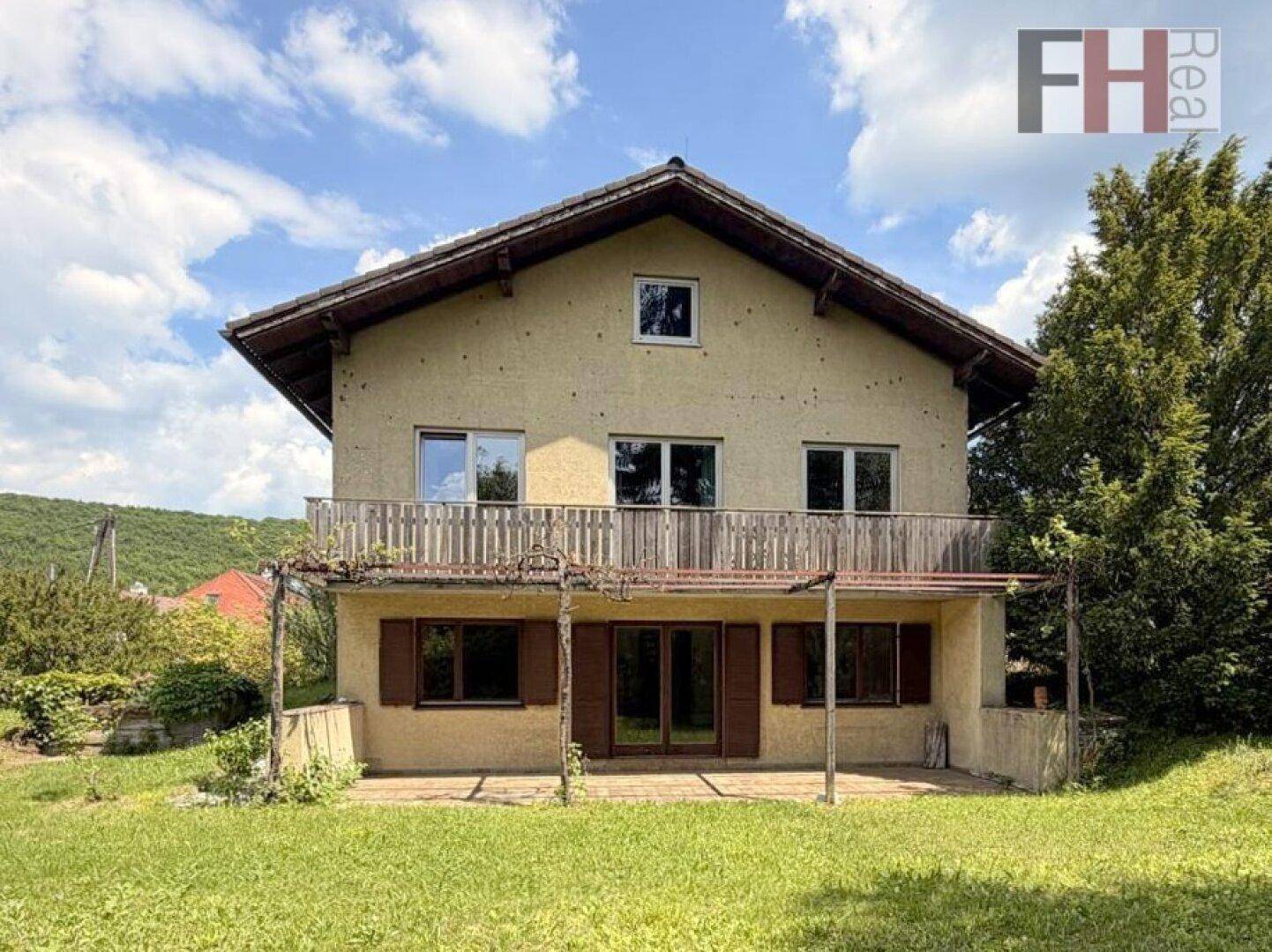 Hausfassade mit Balkon und überdachter Terrasse in ländlicher Umgebung unter blauem Himmel.