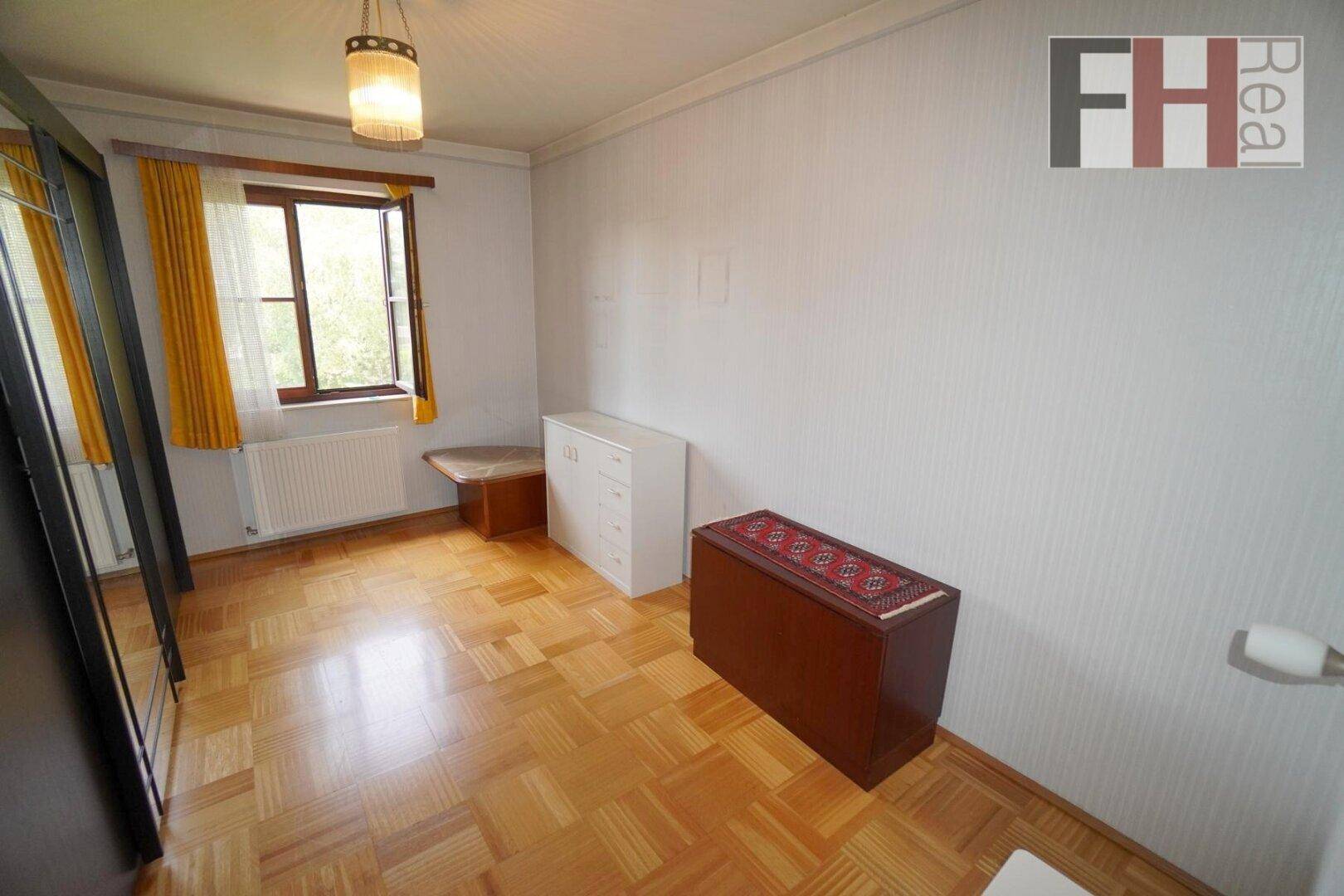 Schlafzimmer mit Parkettboden, Fenster mit gelben Vorhängen und einfacher Einrichtung.