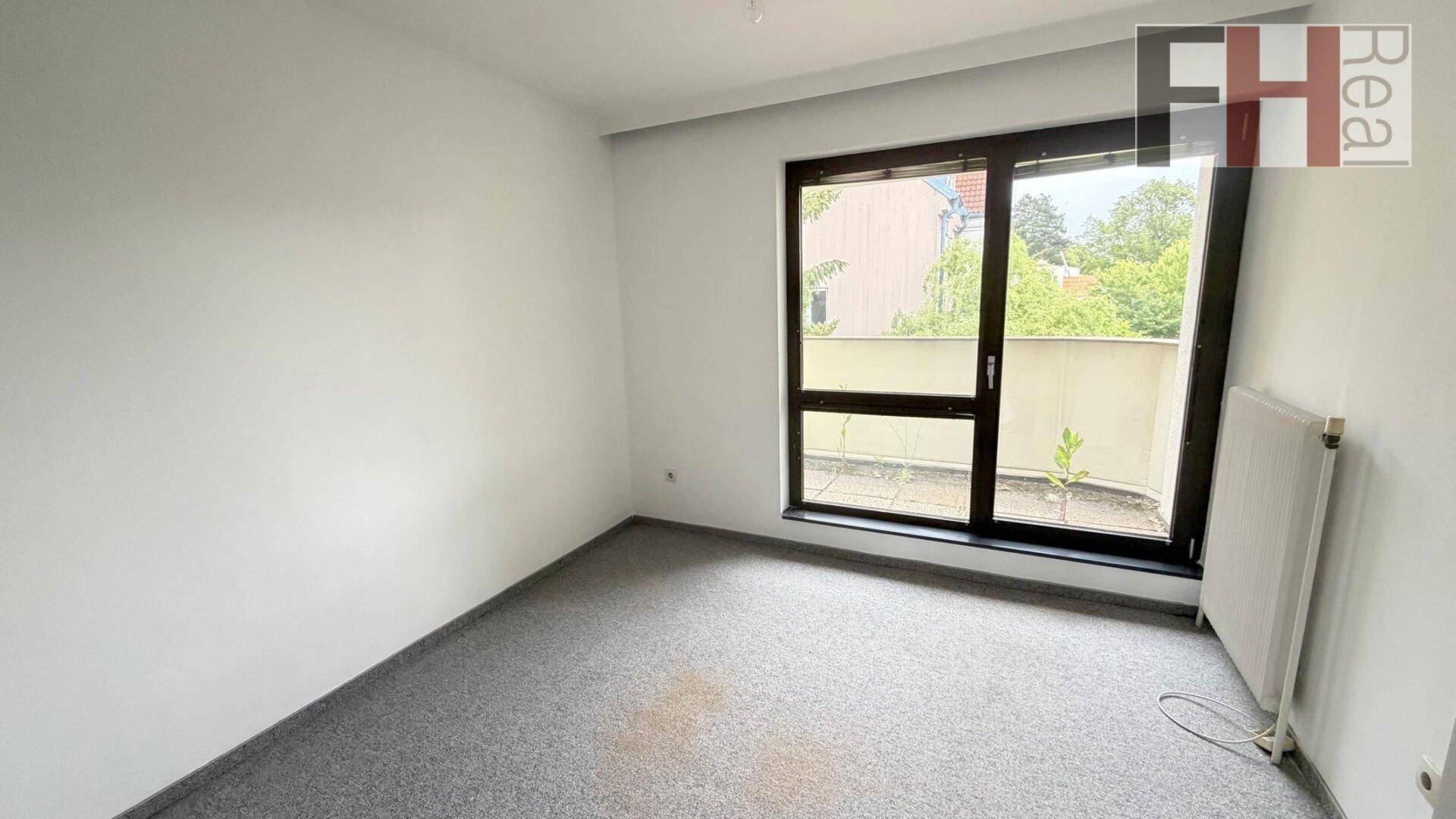 Geräumiges Zimmer mit grauem Teppichboden und großem Fenster mit Zugang zu einem Außenbereich.