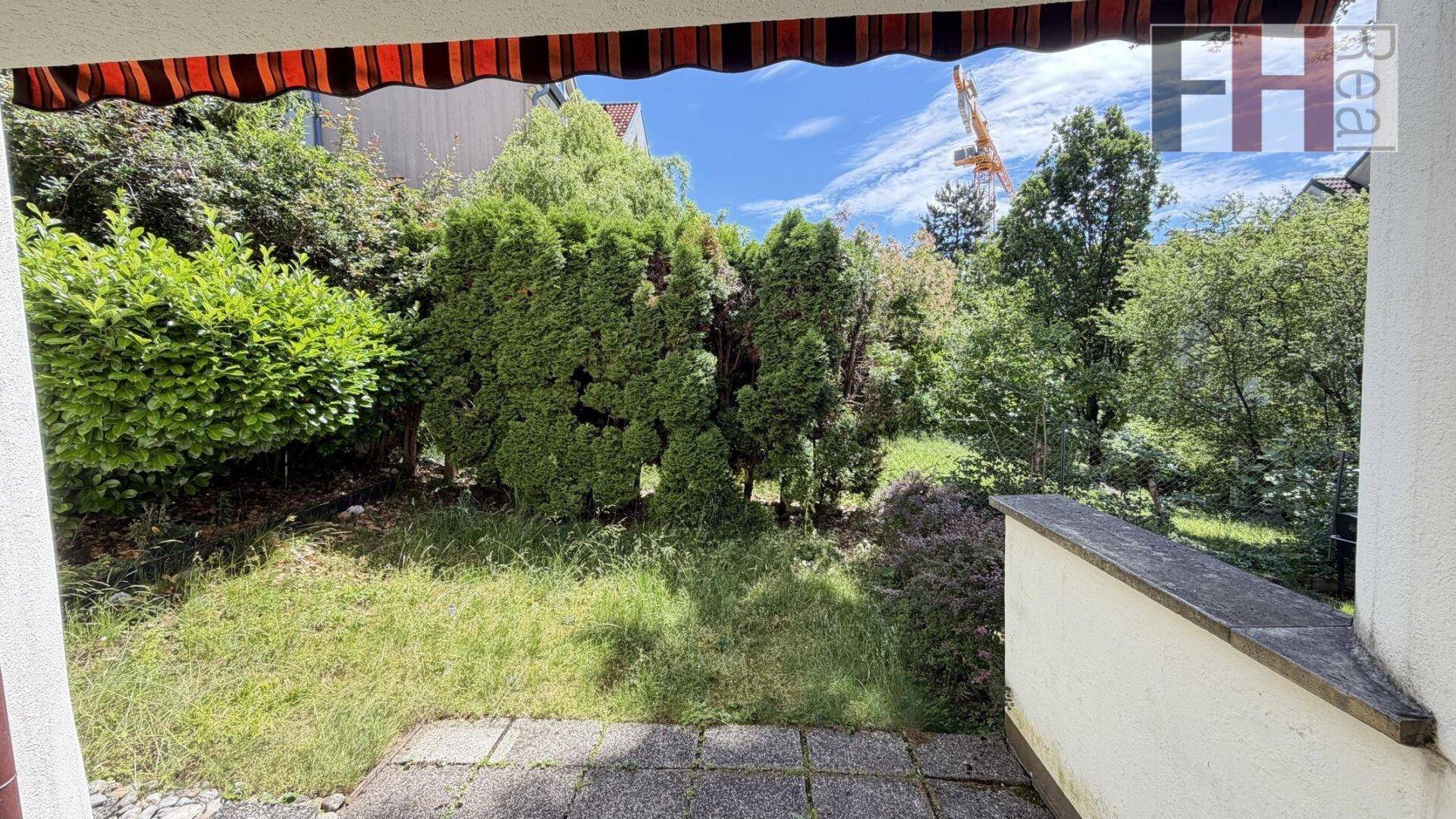 Kleiner Gartenbereich mit Rasen und Sträuchern, geschützt durch eine gestreifte Markise.