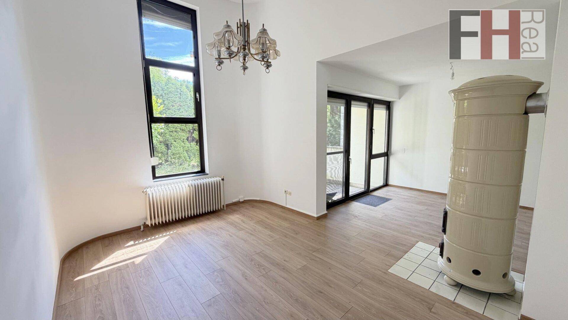 Helles Wohnzimmer mit Laminatboden, großem Fenster, Balkonzugang und einem traditionellen Kachelofen.