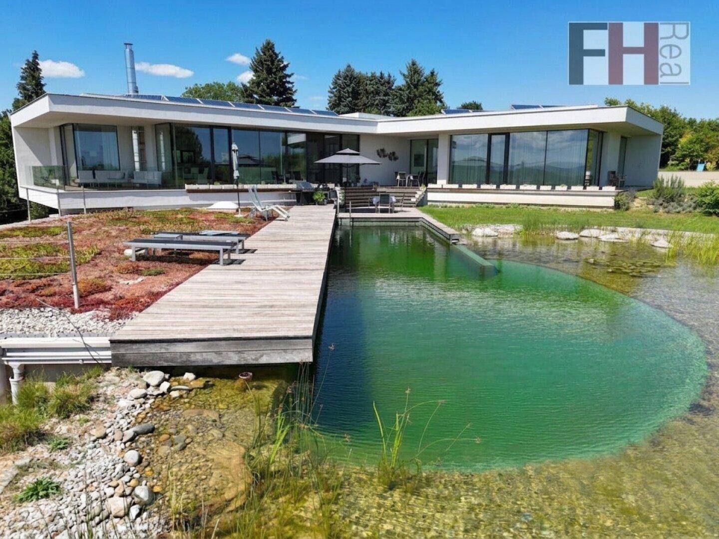 Moderne Villa mit großem Naturpool, Holzdeck und Photovoltaikanlage in grüner Umgebung.
