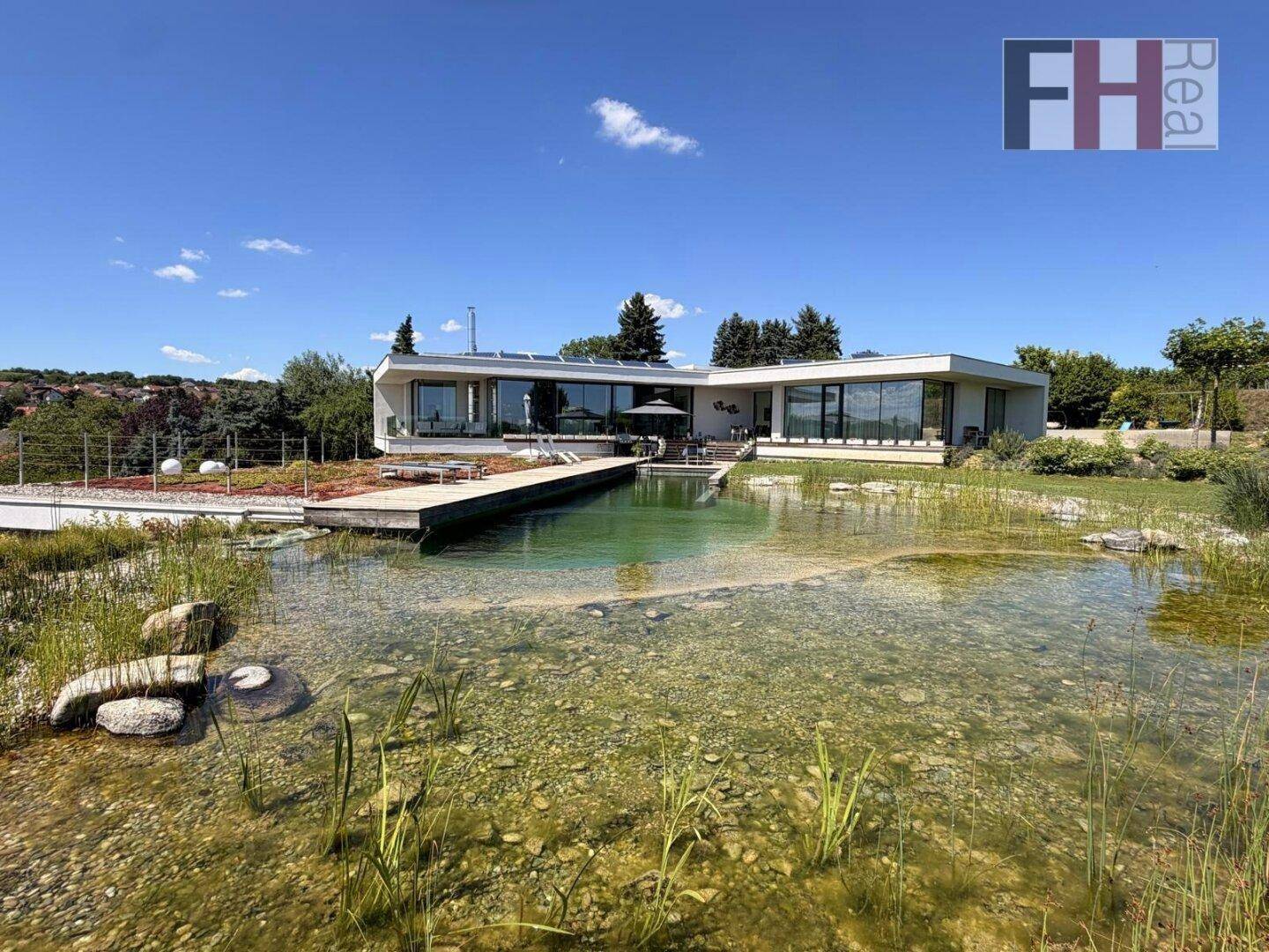 Moderne Villa mit großem Naturschwimmteich und weitläufigem Garten unter blauem Himmel.