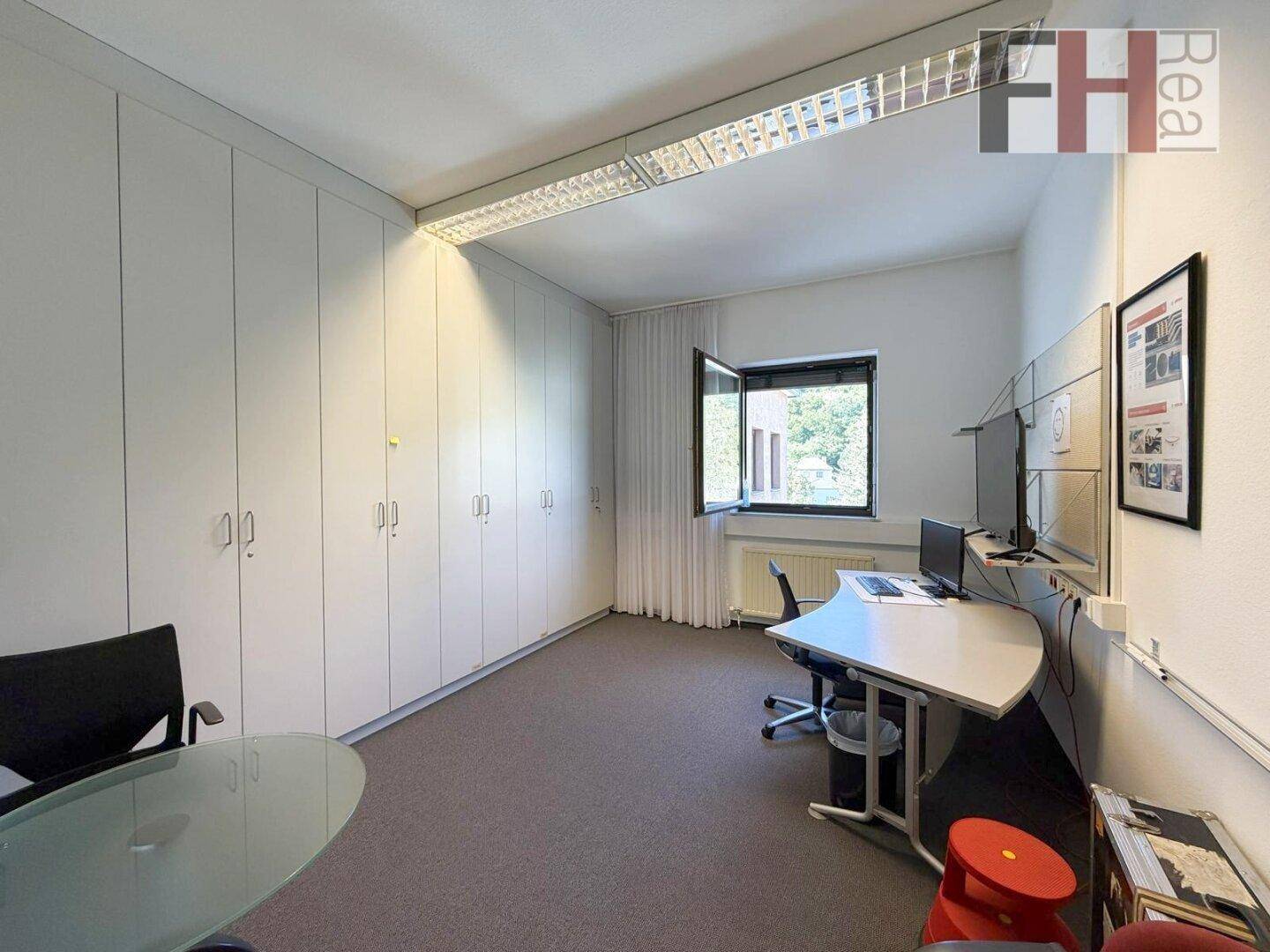 Büro mit einer Wand voller Einbauschränke, Schreibtisch und Fenster mit Blick ins Grüne.