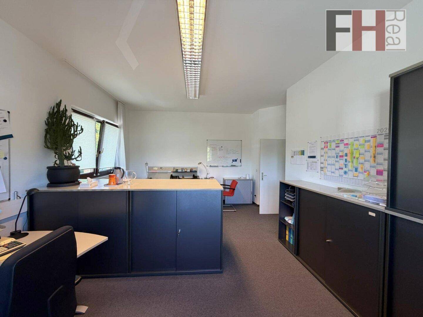 Büro mit mehreren Arbeitsplätzen, dunklen Schränken und einem Fenster, das viel Tageslicht hereinlässt.