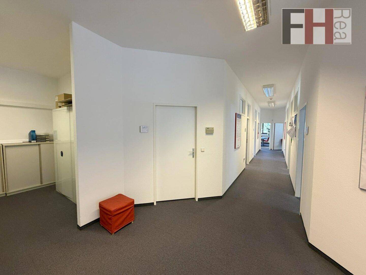 Langer Bürogang mit Teppichboden und weißen Wänden, der zu verschiedenen Räumen führt.