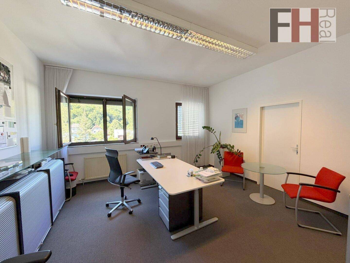 Helles Büro mit Schreibtisch, Bürostuhl und Fenstern, die einen Blick ins Grüne bieten.