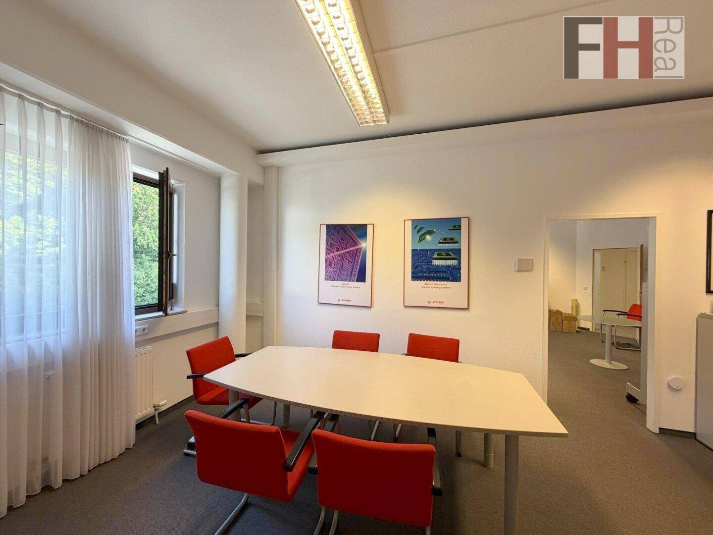 Kleiner Besprechungsraum mit ovalem Tisch, roten Stühlen und einem Fenster.