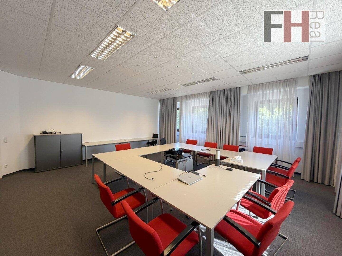 Konferenzraum mit U-förmigem Tisch, Projektor und roten Stühlen für Präsentationen.