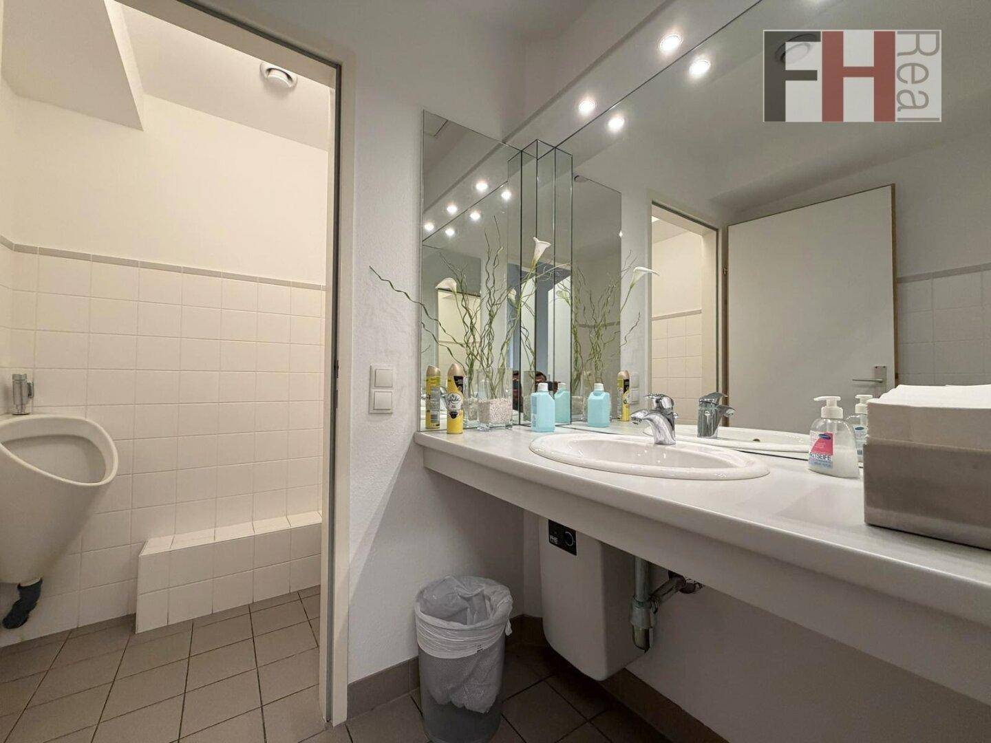 Badezimmer mit Waschbecken, großem Spiegel, Urinal und weißen Fliesen an den Wänden.