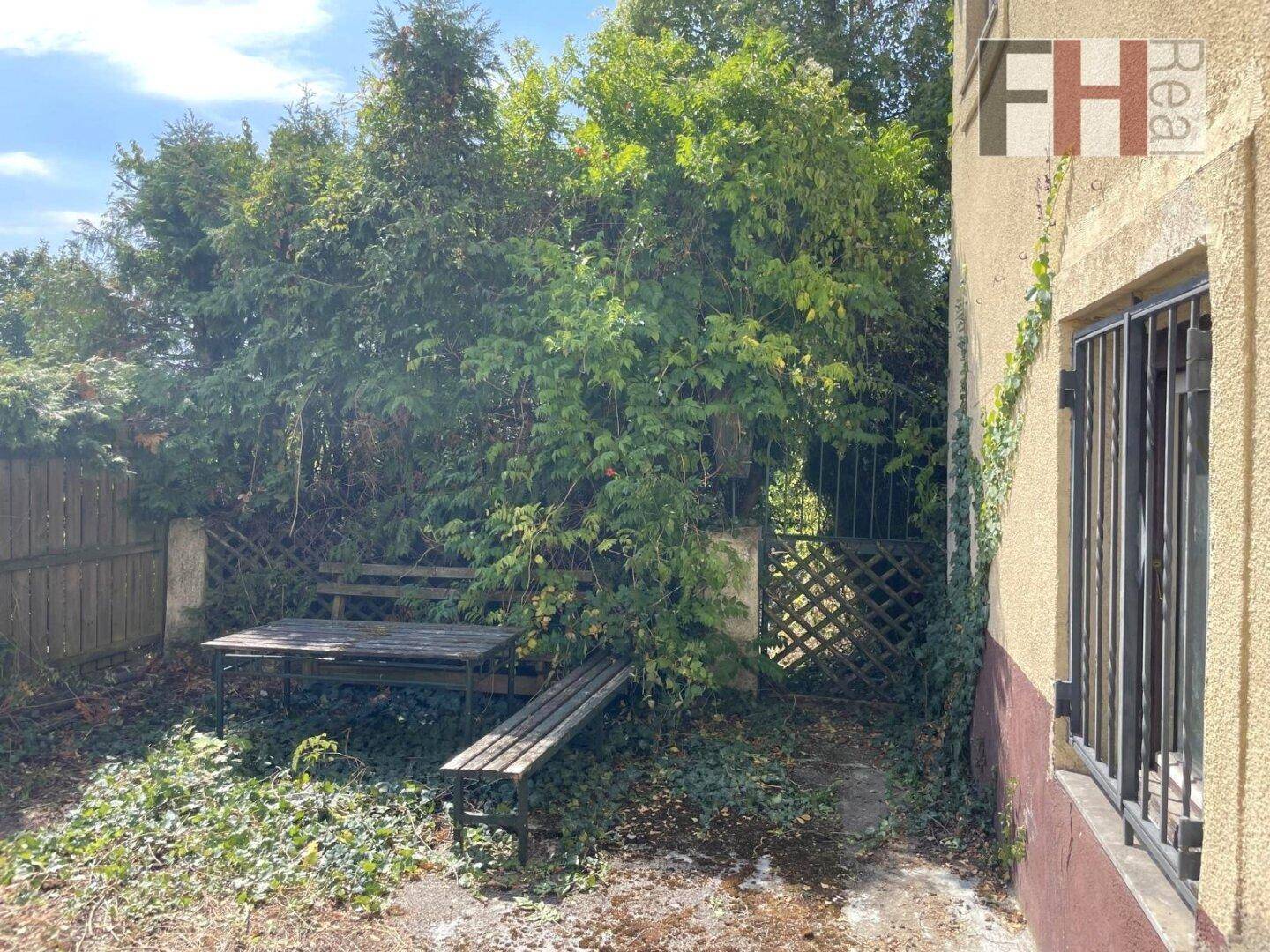 Ein rustikaler Sitzbereich im Freien mit Holztisch und Bänken, umgeben von üppigem Grün und einem Zaun.
