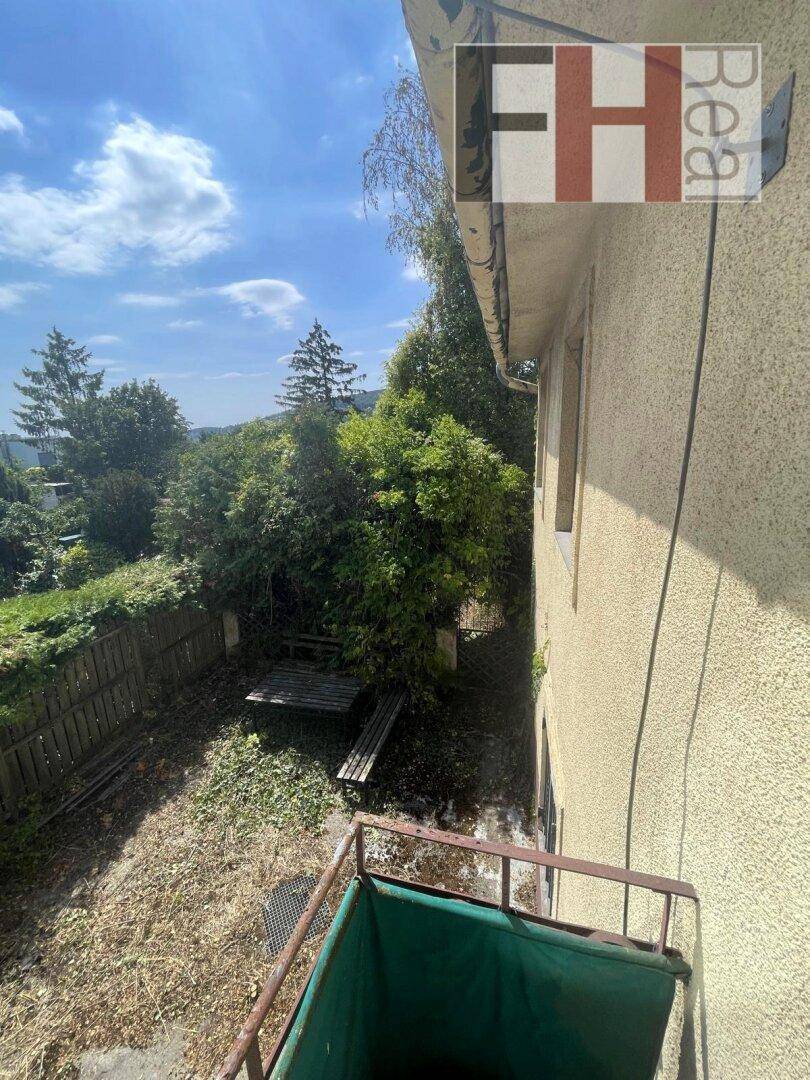 Blick von einem kleinen Balkon auf einen verwilderten Gartenbereich und die Seitenfassade des Hauses.