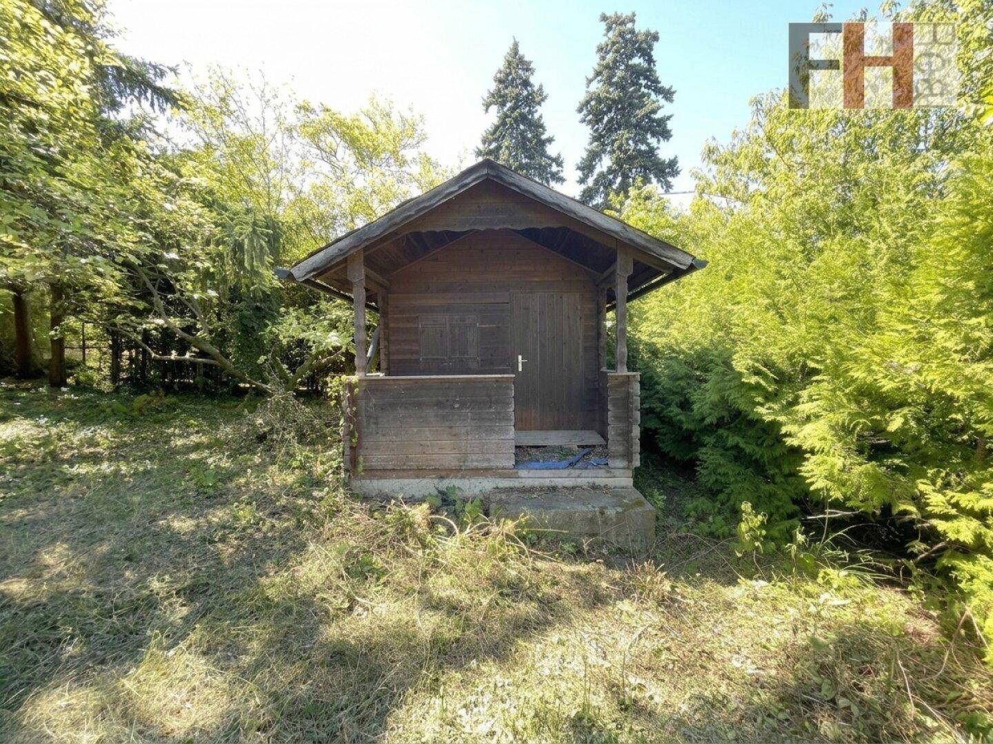 Rustikale Holzhütte mit kleiner Veranda, umgeben von dichtem Grün und hohen Bäumen.