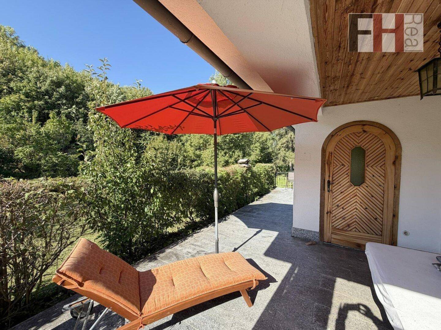 Gemütliche überdachte Terrasse mit Sonnenschirm und Liegestuhl, umgeben von grünen Hecken.