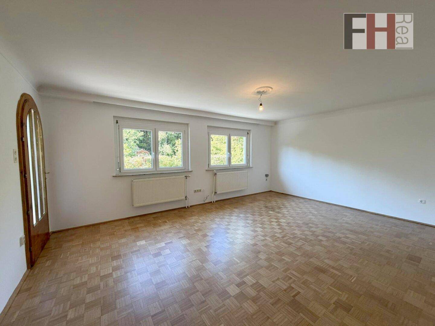 Geräumiges Wohnzimmer mit hellem Parkettboden und großen Fenstern, die viel Licht hereinlassen.