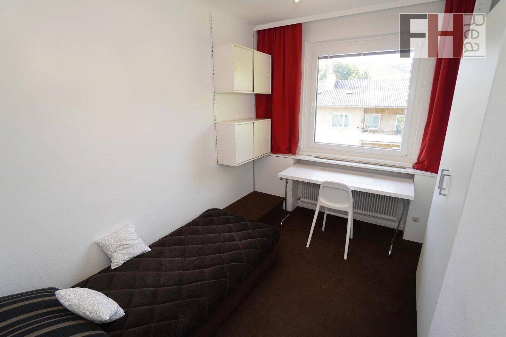 Funktionales Schlafzimmer mit Einzelbett, Schreibtisch und roten Vorhängen am Fenster.