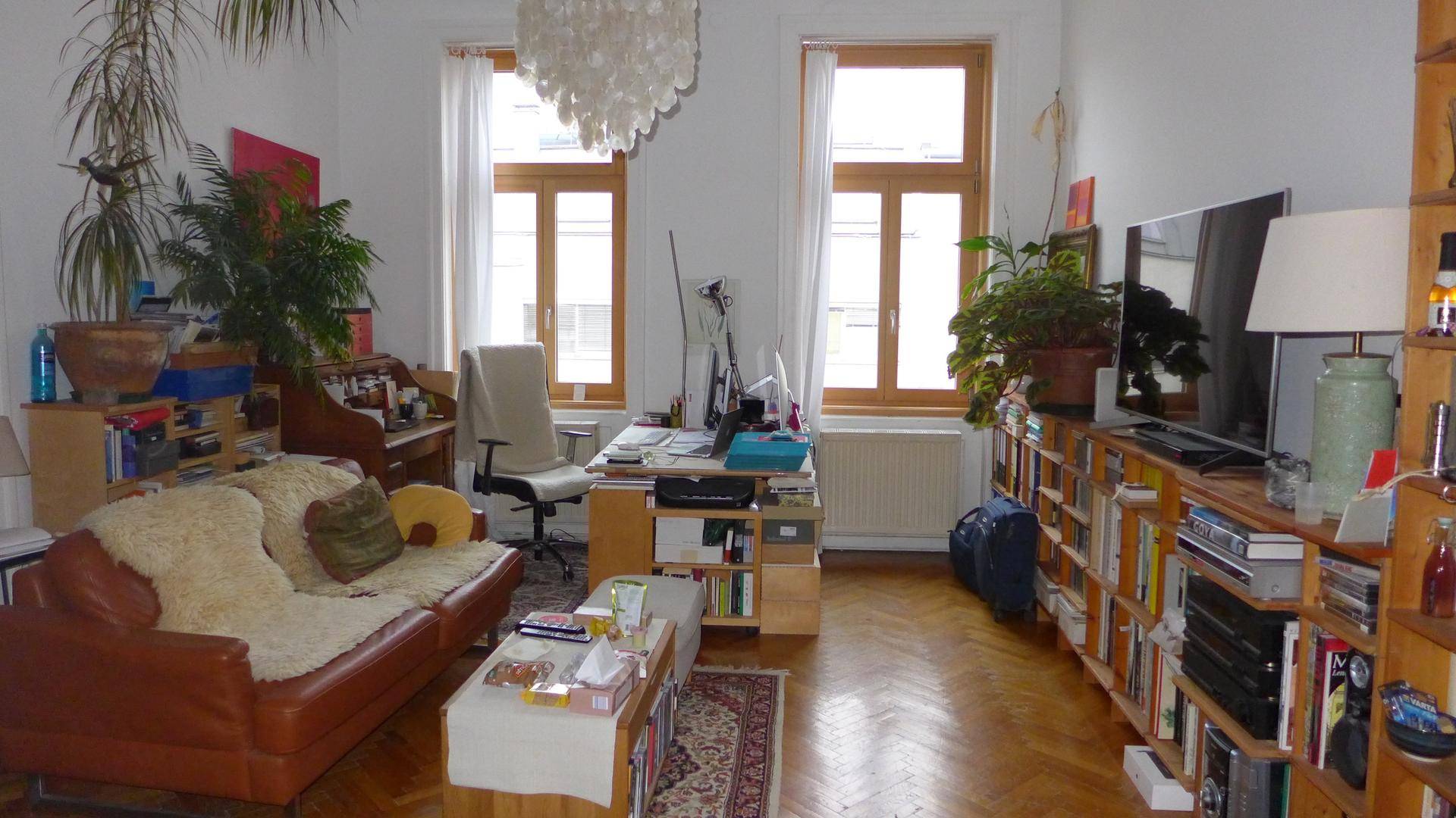 Gemütliches Wohnzimmer mit Ledersofa, Schreibtischbereich, Bücherregalen und großen Fenstern.