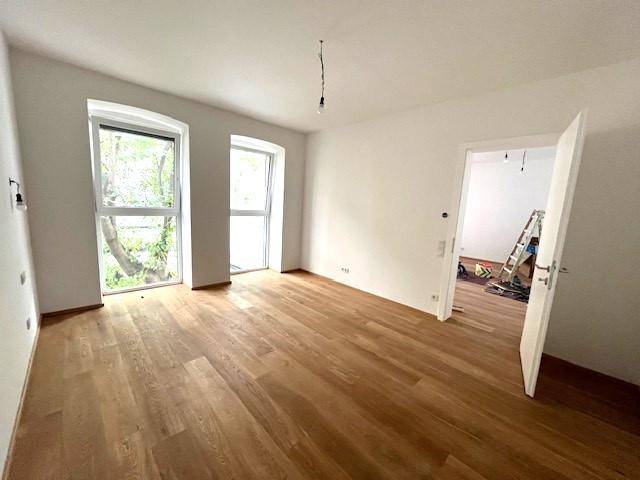 Leeres Zimmer mit Holzboden und zwei Fenstern, die viel Tageslicht hereinlassen.