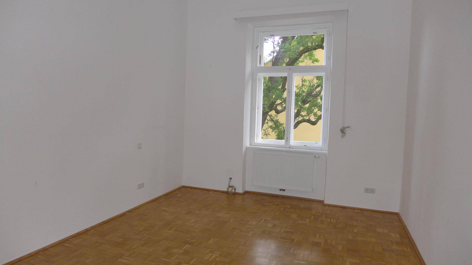 Ruhiges Zimmer mit Parkettboden und einem großen Fenster, das einen Blick auf Bäume bietet.