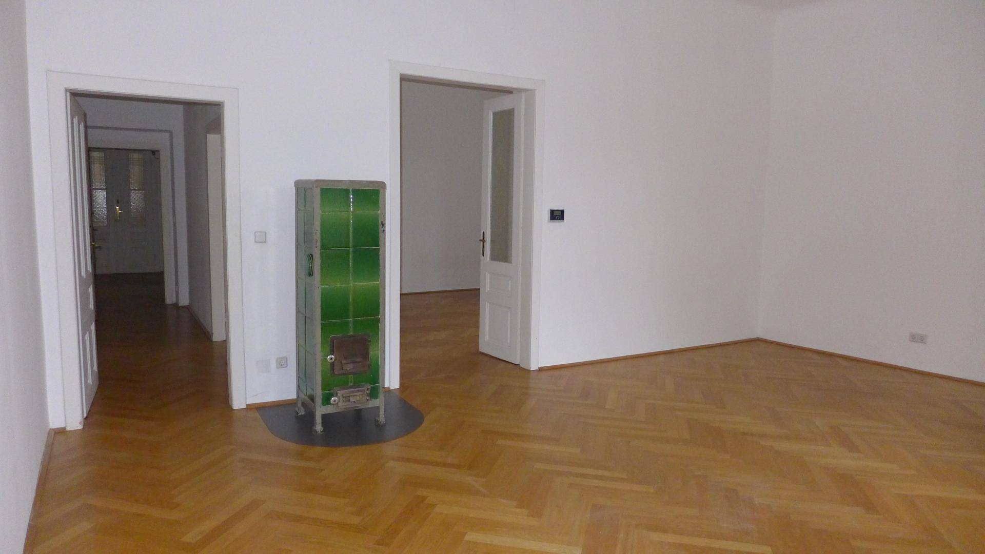 Geräumiges Zimmer mit Fischgrätparkett, einem grünen Kachelofen und Zugang zu weiteren Räumen.