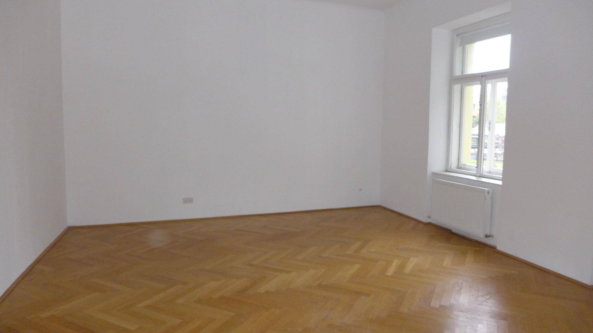 Helles, leeres Zimmer mit Fischgrätparkett und einem großen Fenster, das viel Tageslicht hereinlässt.