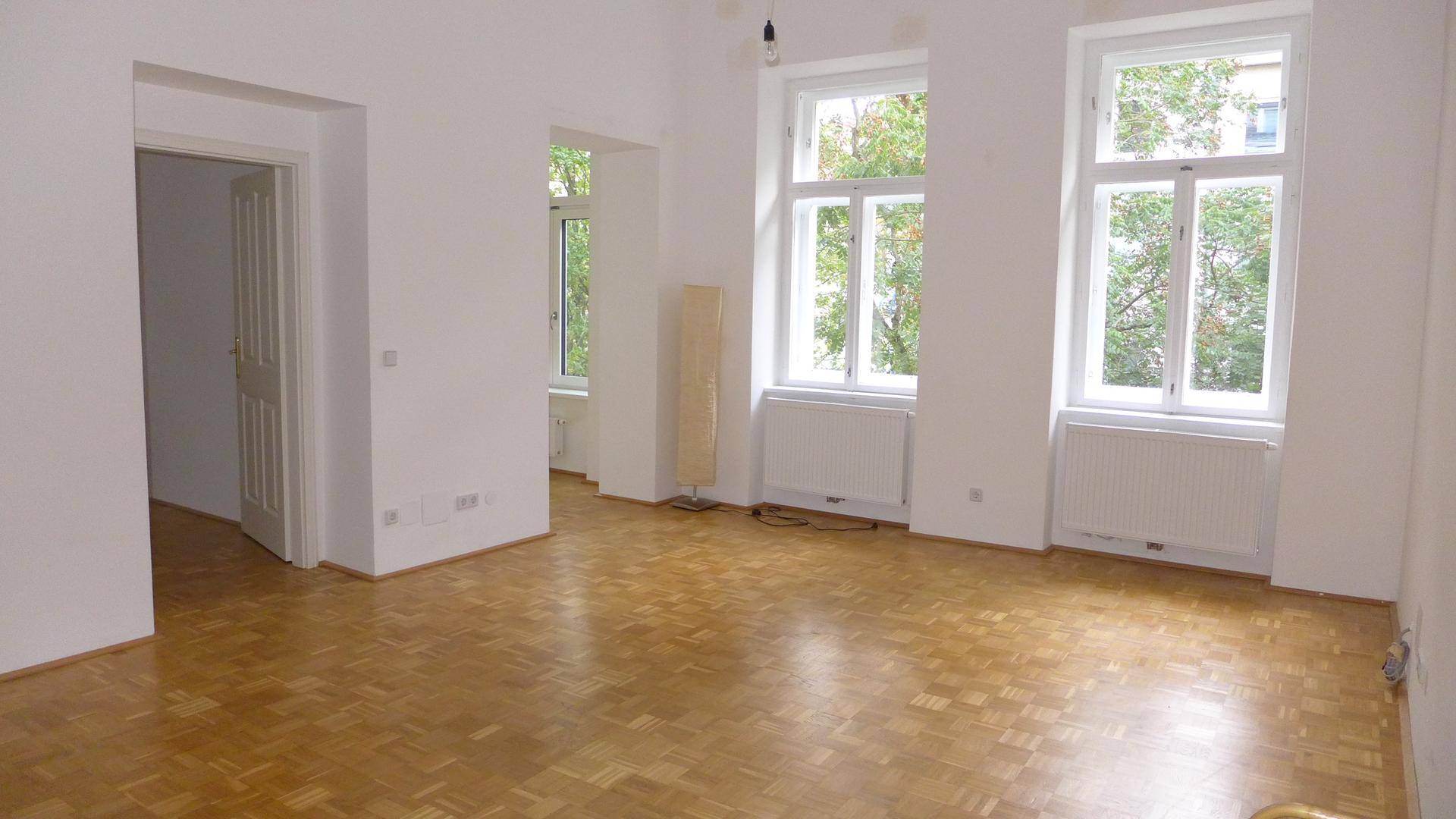 Großzügiges Wohnzimmer mit Parkettboden und zwei hohen Fenstern, die den Raum erhellen.