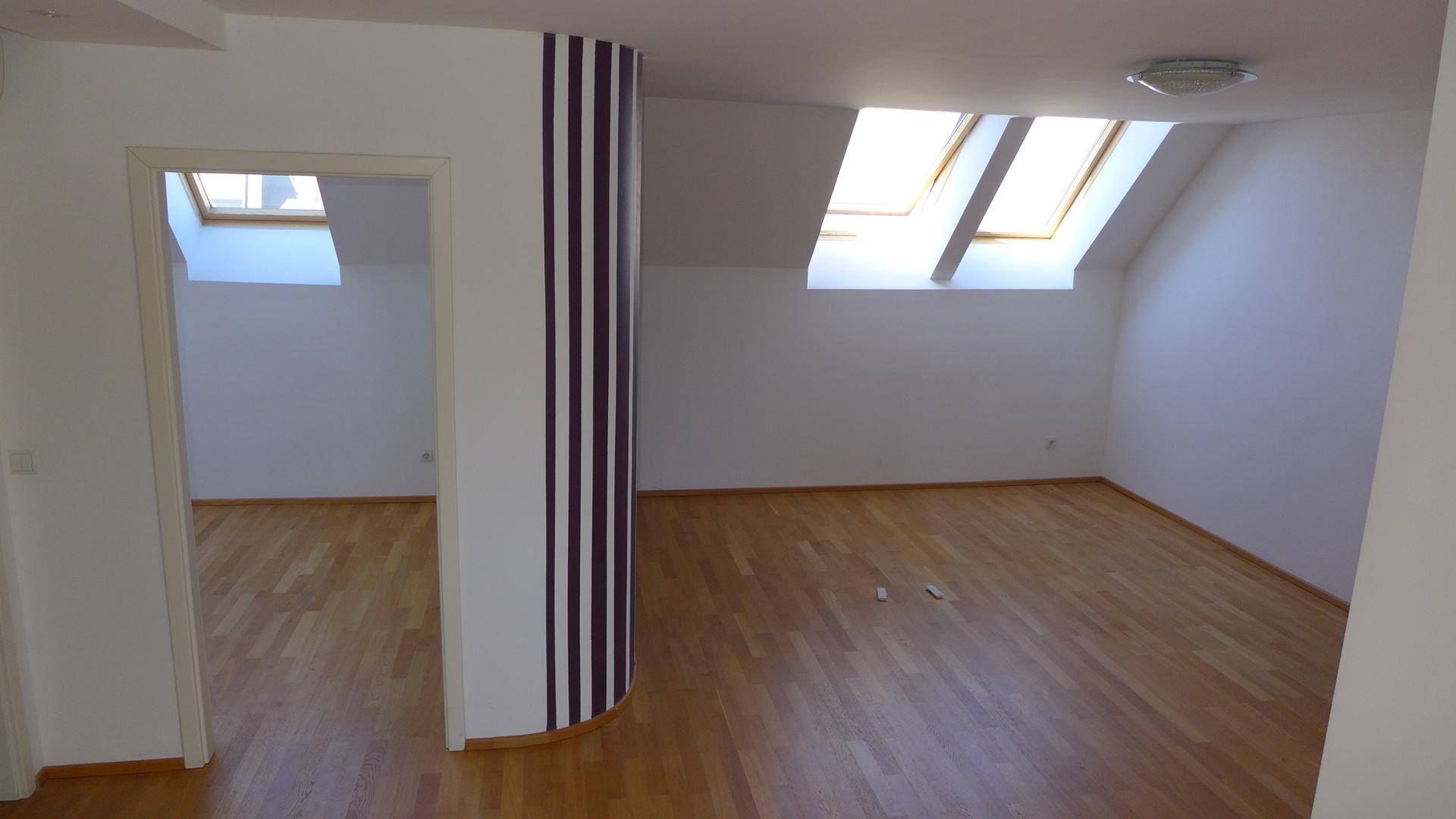 Geräumiger Dachgeschossraum mit Holzparkett, schrägen Wänden und zwei hellen Dachfenstern.