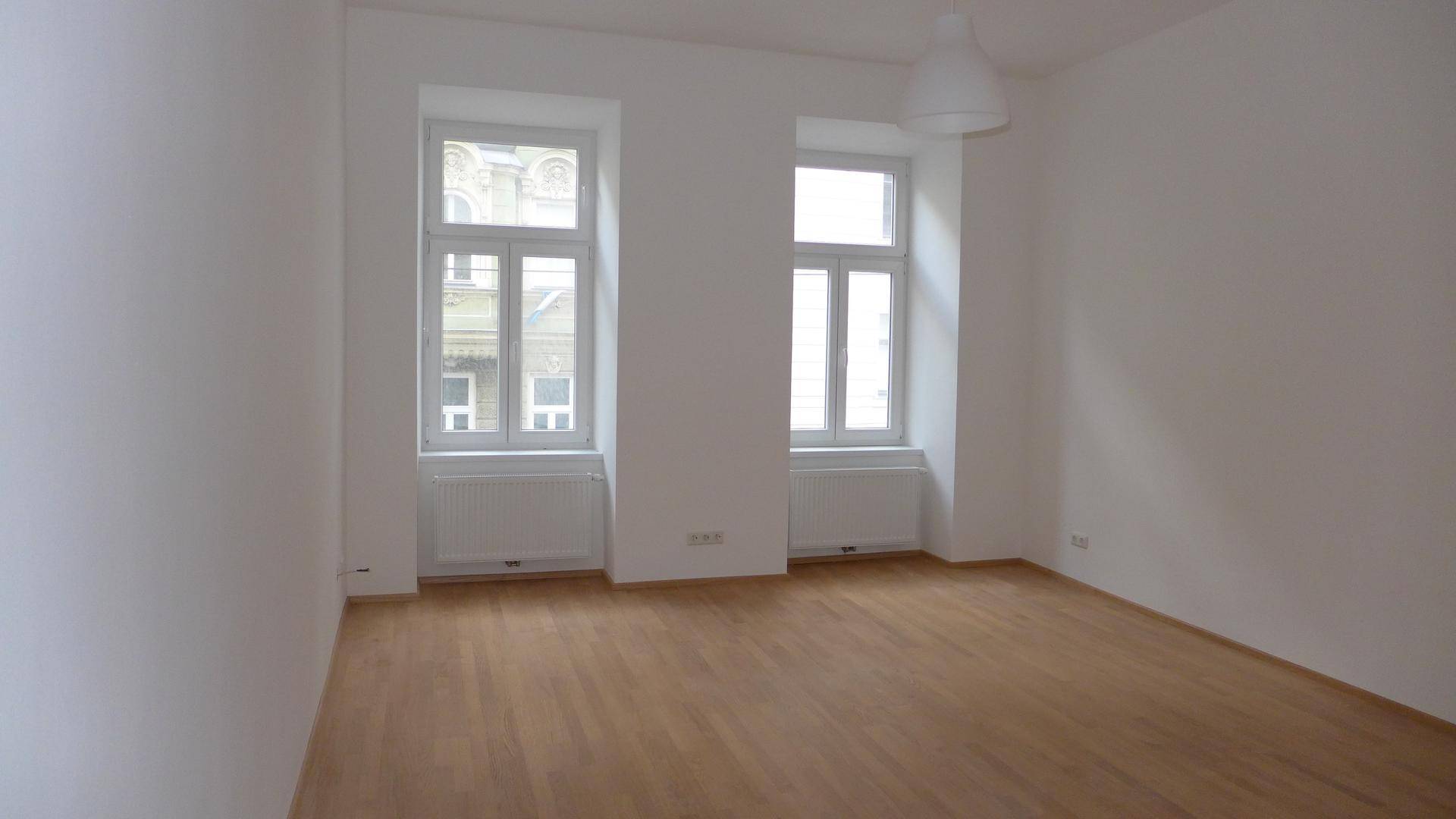 Geräumiges Zimmer mit hellem Parkettboden und zwei großen Fenstern, die viel Tageslicht hereinlassen.