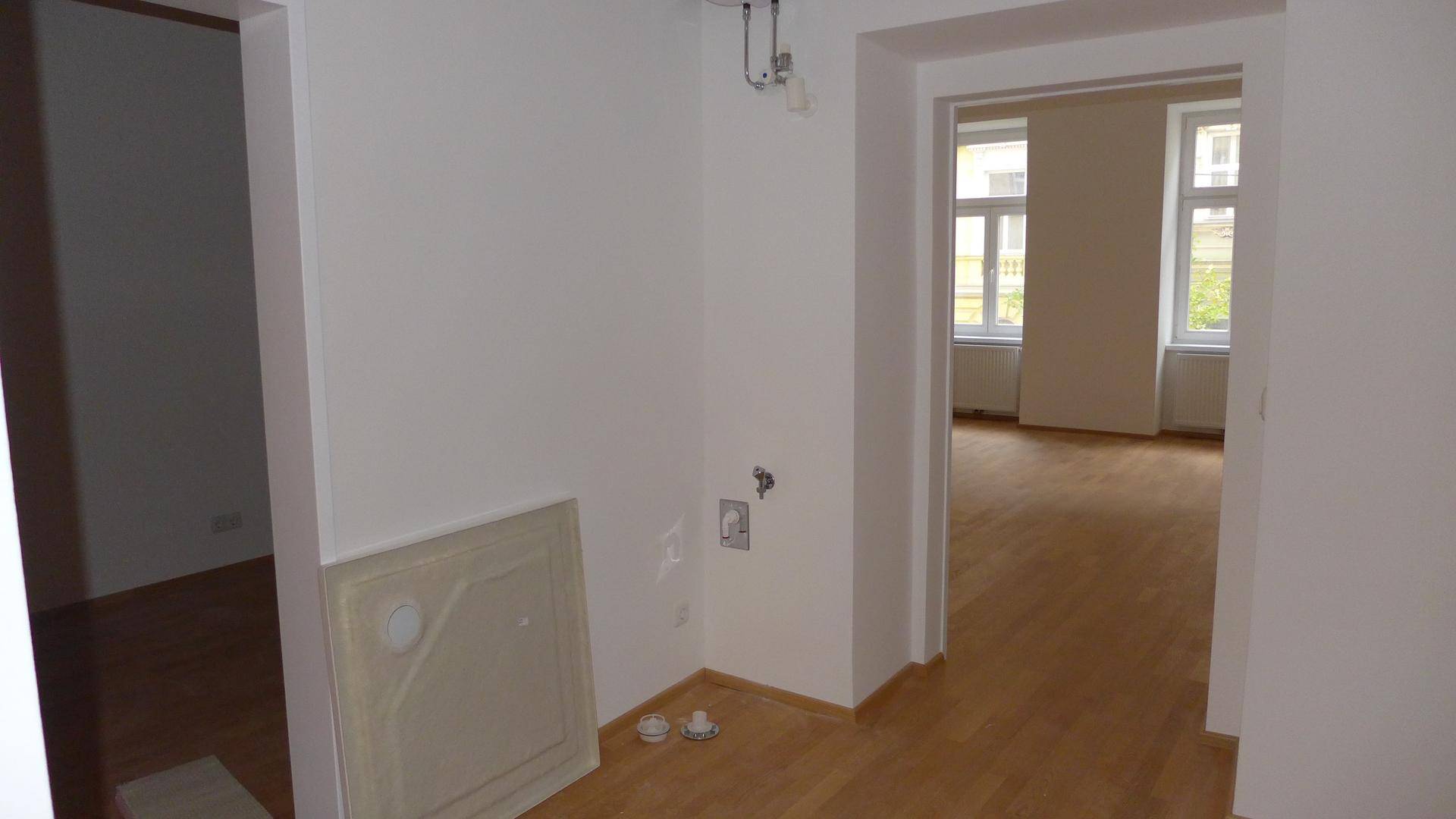 Leerer Küchenbereich mit vorbereiteten Anschlüssen und Zugang zu einem weiteren hellen Zimmer.