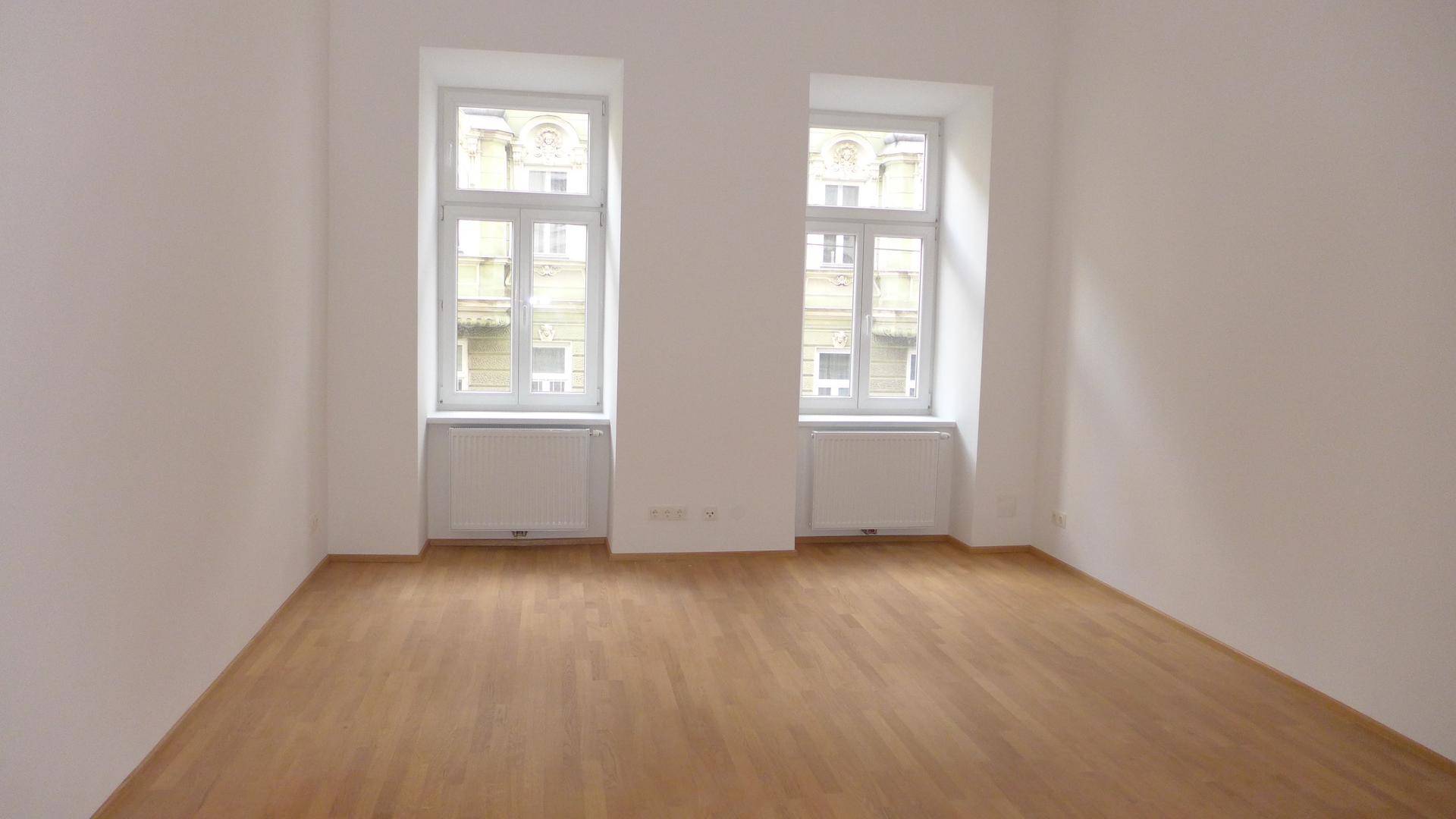 Lichtdurchflutetes Zimmer mit Holzboden und zwei Fenstern, die einen Blick auf die Stadtarchitektur ermöglichen.